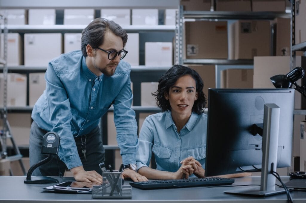 Male and Female Warehouse Inventory Managers Talking, Solving Problems, Using Personal Computer and Checking Stock. In the Background Rows of Shelves Full of Cardboard Box Packages.